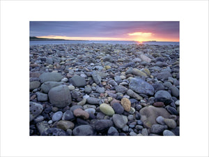 A view of a pebble beach towards the sea, the pebbles are a variety of colours and the horizon is a bright red orange glow