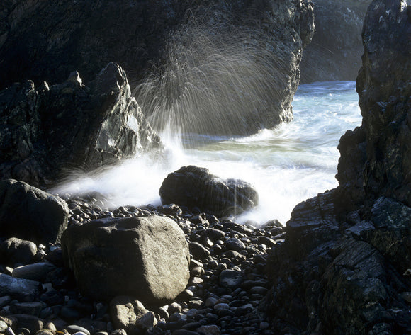 A view of Kynance Cove between the rocks, the water spills over pebbles and the spray is caught by the morning sunlight