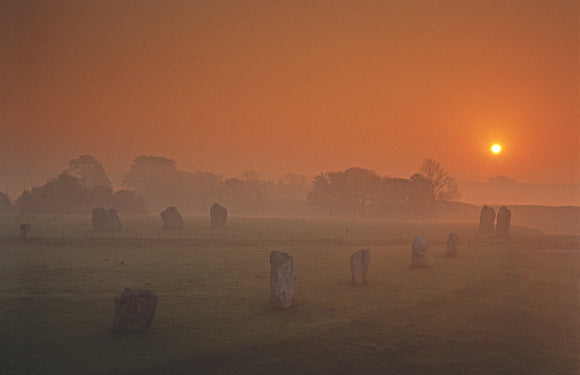 Dawn at Avebury Stone Circle with two stones against a misty landscape with the sun rising in a pink-orange sky