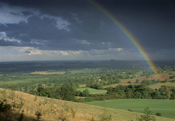 A long distance view over the sun bathed fields of the Clent Hills, Hereford & Worcester, to the dark stormy sky on the horizon with a rainbow streaking across it
