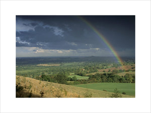 A long distance view over the sun bathed fields of the Clent Hills, Hereford & Worcester, to the dark stormy sky on the horizon with a rainbow streaking across it